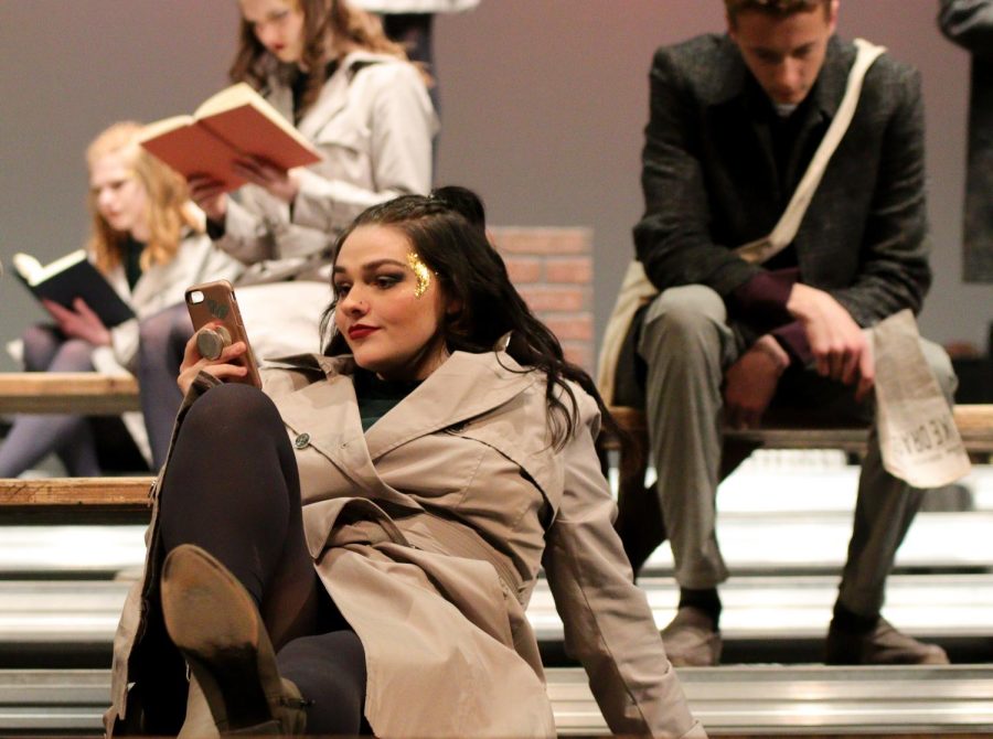 Caily Jones sits on the stage and pretends to scroll through her phone. Her character is waiting for the bus to arrive. 